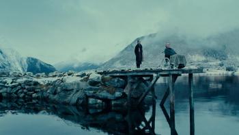 Fjord: A woman stands behind a man, who sits in a wheelchair at the end of a peer, overlooking a lake surrounded by snowy mountains