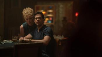 Night Always Comes: A man looks at someone off camera as he sits at a table, a woman standing behind him