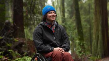 Still from Full Circle: White professional sit skier Trevor Kennison sits in a wheelchair in the woods, wearing a blue ski cap, a red, white, and blue fleece sports shirt, black Eddie Bauer windbreaker jacket, brown pants, and black sport shoes