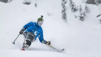 Still from Full Circle: A sit skier heads down a snow-covered mountain, wearing a blue ski outwear jack, green helmet with a GoPro camera on top, and a black sit ski featuring the name Eddie Bauer on display in white letters