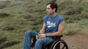 Still from Full Circle: White alpinist and author Barry Corbet sits in a wheelchair on a mountain while holding a green bottle, wearing blue jeans, a blue T-shirt with the name Maui in white letters, and brown sneakers