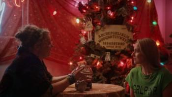 Ghosts of Christmas Past: A young White blond woman sits in a festive tent in front of a fortune teller with a snow globe for a crystal ball.