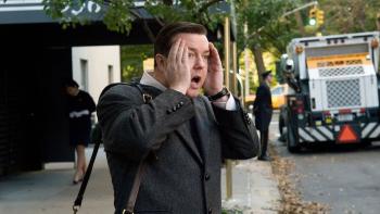 Ricky Gervais stands on a city sidewalk in a suit, hands pressed to the sides of his face in shock.