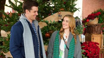 Ghosts of Christmas Past: A young White blond woman and a young White man with black hair, walk together in front of a Christmas tree, smiling.