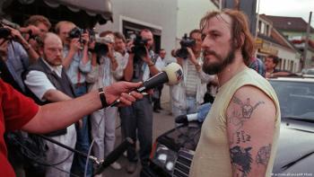 A man with a tattooed arm speaks into a microphone in front of a group of reporters.