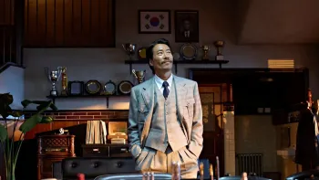 Good News movie: A Korean man in a light grey business suit center stands tall in his fancy office, awards and trophies line the wall behind him