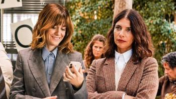 Go Team! Movie: Two women in business attire, one smiling at a phone, the other with crossed arms.