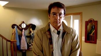 John Cusack stands in the entryway of a family house. He wears a collared shirt, tan jacket, and glasses.