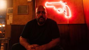 Guns Up movie: Kevin James sits in a nightclub with a red neon pistol on the wall behind him