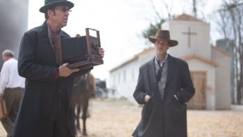 Gunslingers Movie: Ben (Nicolas Cage), wearing a hat and dark glasses, holds an old-fashioned camera; another man watches him