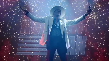 The Running Man: Colman Domingo in white suit and hat stands with arms outstretched on stage amid falling confetti and dramatic lighting