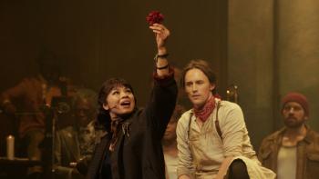 Hadestown the Musical: Eurydice (Eva Noblezada) holds a red flower up and sings while Orpheus (Reeve Carney) looks on