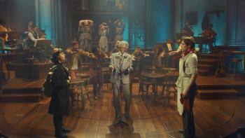 Hadestown the Musical: Eurydice (Eva Noblezada) and Orpheus stand across from each other, lovestruck, while Hermes André De Shields sings between them
