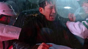 Hard Days movie: Japanese man in black coat center struggles with Japanese police in white uniforms left and right in the rain in front of a car
