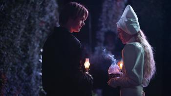 100 Nights of Hero: A young White man and woman stand in the dark holding lanterns, surrounded by tall hedges