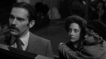 Hester Street: Steven Keats speaks to someone behind a high desk, out of shot, as Carol Kane looks on from behind, holding their young son