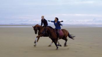 Of Girls and Horses Movie: Two people riding horses on a beach, arms outstretched, cloudy sky.