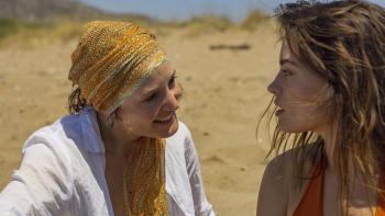 Hot Milk: A young White woman smiles as she looks at another young White woman, the beach in the background