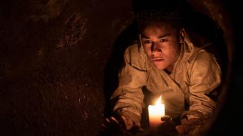 House of Spoils movie: Black woman in dirty white chef's coat center right, crawls through dirt underground tunnel with candle in hand