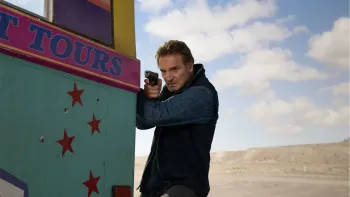 Ice Road: Vengeance movie: Liam Neeson center holds onto the back of a bus while pointing a gun at his target, blue sky, clouds, and desert behind him