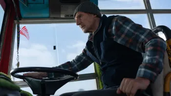 Ice Road: Vengeance movie: Liam Neeson center right drives a bus with his hands on the wheel and the gear stick, blue sky and clouds behind him