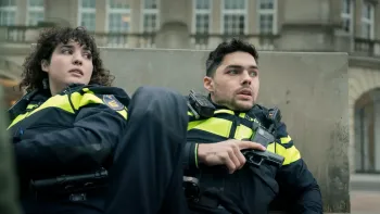 iHostage movie: White Dutch woman police officer left and White Dutch man right both in yellow striped vests lean up against a cement pylon outside