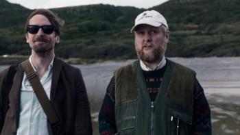 The Ballad of Wallis Island: Two White men, fully clothed, stand on the beach