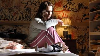 In Her Skin: A young White woman sits on a bed looking sad, in a messy room with writing scrawled on the walls