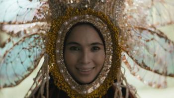 In My Mother's Skin movie: Filipino woman center dressed elaborately in bejeweled mask and headdress that looks like a flower surrounding her face smiles in front of window