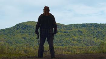 In a Violent Nature Movie: A masked killer stands, back to viewers, at the edge of a ravine, a hook and chain in his hands