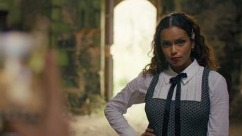 Influencers movie: Georgina Campbell in a white shirt, black ribbon tie, and overalls, right, stands in front of a doorway while outdoors