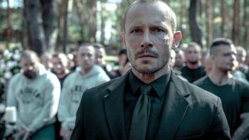 Inside Furioza movie: White Polish bald man with goatee in black suit sits at a funeral, many white bald and short haired men sit behind him