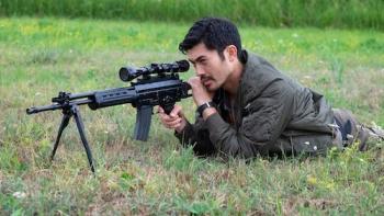A young Asian man lies on the grass taking aim with a sniper rifle