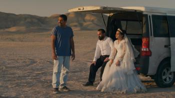 It Was Just an Accident: A group of people, one in a wedding dress, sit in open tailgate of a van in the desert