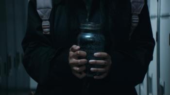 It Lives Inside Movie: A figure wearing black and the straps of a backpack clutches a jar with white-knuckled fingers
