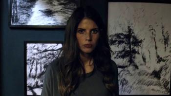 It's Not Over movie: White woman with long brown hair stands in front of three black and white pencil drawings framed on wall