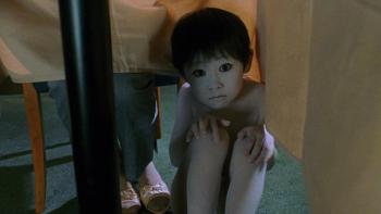 Ju-On—The Grudge: A ghostly child with wide eyes crouches under a table, looking forward