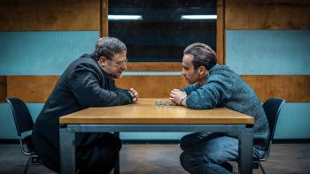 Justice: Two men lean toward each other across a table. One man is handcuffed and chained to the table.