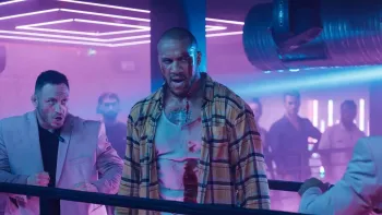 K.O. movie: Black French man, bloodied, faces off against two opponents in white suits in pink and purple lit nightclub, onlookers behind them