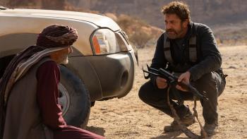 Kandahar Movie: Mo and Tom, who holds a rifle, crouch by the front of a car