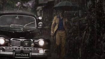 K.G.F -- Chapter 1: An Indian man in a yellow-brown suit holds an umbrella in the rain while next to a black car