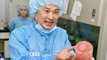 till from King of Clones: South Korean stem cell scientist Huang Woo-suk looks at an interviewer and the camera while pointing to an object during a conversation about his work, wearing light blue medical scrubs that include a hair cap, mask, and body suit