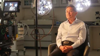 Still from King of Clones: South Korean stem cell scientist Huang Woo-suk seated with hands folded inside a laboratory for a firsthand interview in front of a camera, wearing a white Oxford shirt with the collar opened and brown pants and belt