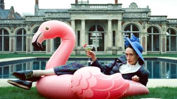 Lady: A White woman with dark brown hair poses on an inflatable flaming, holding a cocktail, in front of the swimming pool of a stately home