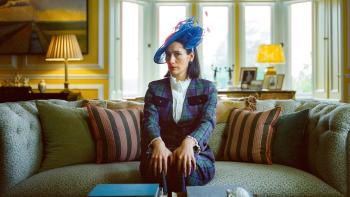 Lady: A White woman with dark brown poses in a suit and formal hat, sitting on a couch in a formal living room