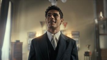 Lefter: The Story of Ordinarius movie: A Greek Turkish man with short black hair and in dark suit and tie, center, stands inside large office