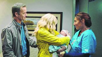 In profile a woman holds an infant while a man behind her and a woman in scrubs look on smiling.
