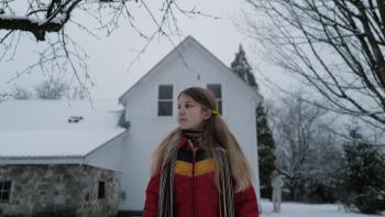 Longlegs Movie: Young Lee Harker stands outside of her house in the snow, a cautious look on her face