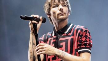 Louis Tomlinson -- All of Those Voices: The head and shoulders of Louis Tomlinson as he looks to his left, holding a microphone