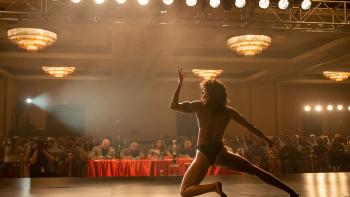 Love Lies Bleeding Movie: A muscular woman wearing a bikini on a stage poses in front of a table of judges in an auditorium